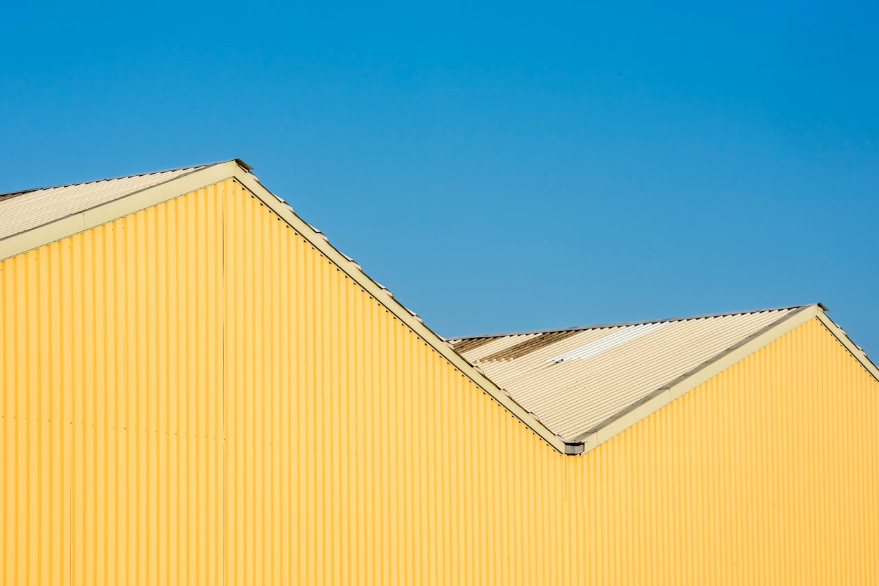 A bold geometric composition of a yellow industrial building roof set against a clear blue sky.